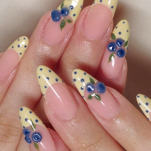 Puede incluir: Primer plano de uñas manicuradas con diseño de manicura francesa. Las uñas están pintadas con una base rosa claro, puntas amarillas con lunares azules y detalles de arándanos y hojas en 3D. Las uñas son largas y almendradas.