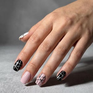 Puede incluir: Una mano con uñas manicuradas en varios diseños. Algunas uñas son negras con patrones geométricos blancos, otras son de color rosa pálido y una es blanca con un diseño similar. Las uñas están sobre un fondo gris.