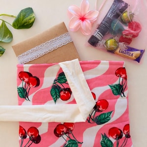 Puede incluir: Una bolsa a rayas rosas y blancas con un estampado de cerezas, un sobre marrón con ribete de encaje, una bolsa de organza rosa con caramelos y un tubo de producto, y una flor rosa.