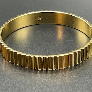 May include: A gold-colored bangle bracelet with a ridged, vertical design around the outer edge. The bracelet has a smooth interior and a small marking. The jewelry is displayed on a dark surface.