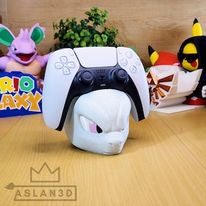Op de afbeelding: Een witte en zwarte PlayStation 5-controller rust op een lichtgrijze Mewtwo-vormige standaard. De standaard heeft roze ogen en een textuur. Andere beeldjes en groen zijn op de achtergrond. Het logo "ASLAN3D" staat onderaan.