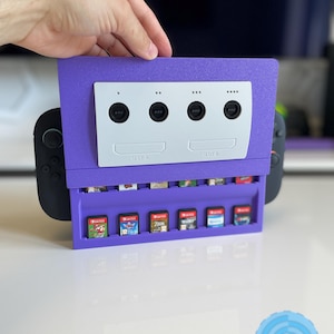 Könnte beinhalten: Ein lila Videospiel-Kassettenhalter mit einer weißen Frontplatte, die einer klassischen Spielkonsole ähnelt. Der Halter zeigt mehrere Spielkassetten in einer Reihe. Der Text "SLOT A" und "SLOT B" sind sichtbar.