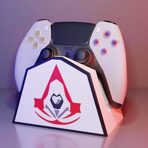 Peut inclure: Une manette PlayStation 5 blanche et noire repose sur un support blanc avec le logo Assassin's Creed en rouge et noir. La manette a un centre noir et des côtés blancs avec des boutons colorés. Le support est incliné et a un design géométrique.
