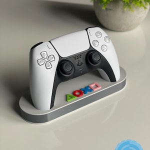 Op de afbeelding: Een wit en zwarte gamecontroller op een grijze standaard met kleurrijke symbolen. De standaard heeft de tekst "AOXO" in groen, rood, blauw en roze. Een kleine groene plant in een grijze pot staat op de achtergrond.