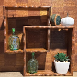 May include: A brown wooden wall shelf with a wavy edge design featuring two interlocking square sections. The shelf holds two green glass bottles with horse imagery, a round vintage tin, a white ceramic pumpkin, and a small green succulent.