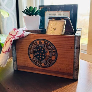 Puede incluir: Una caja de madera con bordes reforzados de metal, con un logotipo en blanco y negro de Procter & Gamble Trade Mark. Dentro de la caja hay un pequeño reloj dorado, una maceta blanca con una suculenta verde y un paño blanco doblado con bordados rojos.