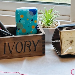 Puede incluir: Una caja de madera con la palabra "IVORY" en letras negras, que contiene bolígrafos y una caja de pañuelos de color turquesa con un patrón de círculos azules y naranjas. Un reloj de viaje vintage dorado está abierto junto a una pequeña planta en maceta.