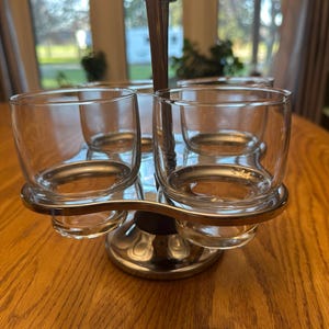 Puede incluir: Un juego de vasos de cóctel vintage con cuatro vasos transparentes y un soporte de metal plateado. Los vasos son cilíndricos y se asientan en soportes circulares. El soporte tiene un asa central y una base redonda.