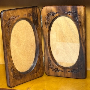 May include: A hinged double photo frame made of dark brown wood with a visible grain. Each rectangular panel features a central oval cutout for a photograph, accented by carved scrollwork patterns in the corners of the wooden frames.