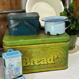 May include: A collection of vintage metal items including a large green Wheat Heart Bread tin, a dark blue rectangular lunch box with a handle, a small light blue enamel mug with rust, a white enamel bowl, and a light blue wall-mounted dispenser.