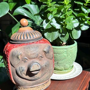 May include: A ceramic pig-shaped jar with a red and brown finish. The jar has a lid with a wooden knob and sits on a metal stand. A green potted plant is in the background.
