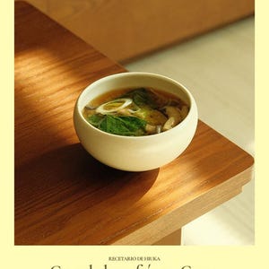 以下が含まれることがあります： 木製のテーブルの上に、緑色の野菜とスライスしたネギが入った白いセラミックボウルに入ったスープ。画像は暖かく、居心地の良い雰囲気で、心安らぐ食事を連想させます。下部のテキストは「Cuando hace frío en Corea」と書かれています。