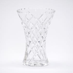 Könnte beinhalten: Eine transparente, geschliffene Glasvase mit ausgestellter Oberseite und Rautenmuster. Die Vase hat eine geschwungene, sanduhrförmige Form und steht auf einem soliden Sockel. Das Glas ist transparent.