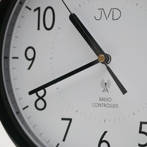 May include: Close-up of a white clock face with black numerals and hands. The clock has a black bezel and the text "JVD" at the top. The words "Radio Controlled" are printed below a radio tower symbol.