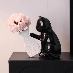 May include: A glossy black ceramic cat figurine holding a white vase with a bouquet of pink flowers. The cat is in a sitting position, with its paw gently touching the vase. The vase has a fish-like shape.