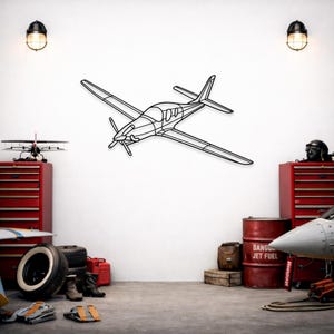 Könnte beinhalten: Ein schwarzes Wandkunstobjekt aus Metall in Form eines einmotorigen Propellerflugzeugs. Das Design zeigt eine minimalistische Umrisszeichnung des Flugzeugs mit markantem Propeller, Tragflächen und Leitwerk, mittig an einer weißen Wand.