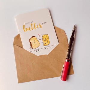 May include: A greeting card with the text "You're my butter half!" is inside a brown envelope. The card features illustrations of a piece of toast and a stick of butter. A red and black fountain pen rests on the white surface.