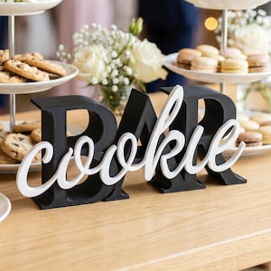 May include: A table laden with treats, including cookies and macarons, presented on tiered stands. White roses and gypsophila decorate the stands. A black and white sign spells out "Cookie Bar".