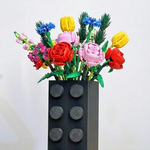 Puede incluir: Un jarrón negro con diseño de ladrillo Lego sostiene un colorido ramo de flores. Las flores son de ladrillos Lego en tonos rojos, rosas, amarillos y azules, con tallos y hojas verdes. El jarrón es rectangular.