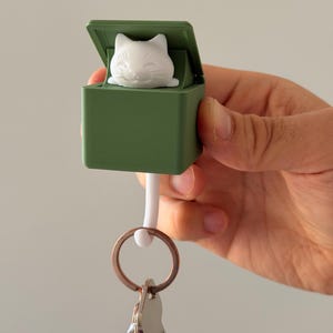 May include: A green key holder featuring a white cat figurine. The key holder is connected to a keyring with multiple keys. The cat figurine is white and has a smiling expression. The key holder is held in a person's hand.