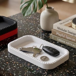 Puede incluir: Una bandeja rectangular blanca con borde de cuentas contiene llaves, un mando a distancia de coche y una moneda. Una bandeja negra y roja está en el fondo. También se ven libros con títulos como "Modular Design" y una lámpara.