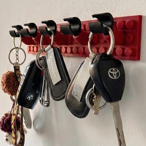 Puede incluir: Un llavero rojo y negro montado en una pared blanca. El llavero está hecho de bloques rojos con ganchos negros, que sostienen varios juegos de llaves con llaveros. Las llaves cuelgan en fila, mostrando una solución de almacenamiento práctica y organizada.