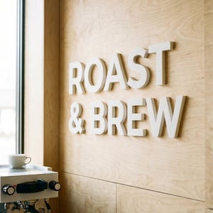 May include: White 3D letters spell "ROAST & BREW" on a light wood paneled wall. A white espresso machine and a small white cup are visible in the lower left corner, suggesting a coffee shop setting.