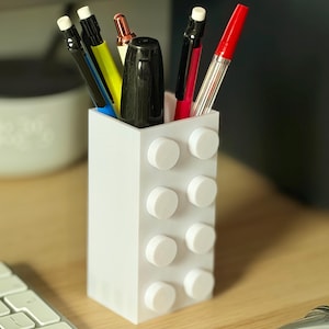 Puede incluir: Un portalápices blanco diseñado para parecer un bloque de construcción, lleno de varios bolígrafos y lápices. Los bolígrafos incluyen bolígrafos negros, rojos y amarillos. El soporte está sobre un escritorio de madera junto a un teclado y un ratón.