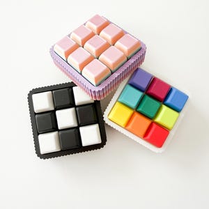 May include: Three square trays of colorful, square-shaped objects. One tray holds pink objects, another black and white, and the third features a rainbow of colors. The trays have decorative edges and are set against a white background.