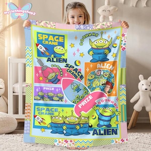 May include: A light blue blanket featuring cartoon aliens and the text "SPACE CRANE" and "ALIEN". The blanket has a patchwork design with colorful borders and illustrations of rockets and stars. The blanket is held by a young girl.