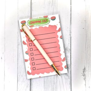 May include: A notepad with a pink shopping list design and a matching pen. The notepad has a scalloped edge and is decorated with watermelon slices. The pen is white with gold accents. The notepad has the words "SHOPPING LIST" at the top.