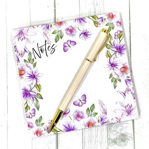 May include: A cream-colored pen with gold accents rests on a floral notepad. The notepad features a white background with purple and pink flowers, green leaves, and butterflies. The word "Notes" is written in black script.