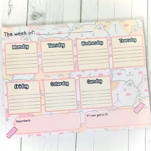 May include: A weekly planner with a cat-themed design. The planner features spaces for each day of the week, with the days of the week written in a playful font. The background is decorated with a repeating pattern of cute cat faces in pastel colors.