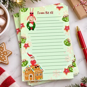 May include: A stack of light green stationery paper featuring the text From the elf at the top. The design includes illustrations of an elf hanging from a wreath, an elf sitting on a gingerbread house, green mugs of cocoa, and red star cookies.