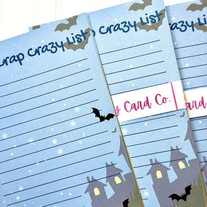 May include: Three light blue lined notepads featuring a dark blue header that reads Bat-Crap Crazy List. The design includes dark silhouettes of bats, a starry night sky, and a haunted house at the bottom. A white band with the text Card Co. wraps around the pads.