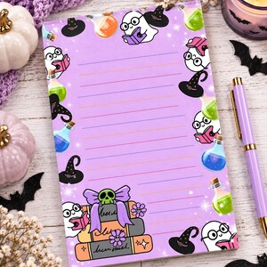 May include: A rectangular notepad with a light purple background featuring horizontal lines. The border is decorated with illustrations of white ghosts wearing glasses and reading books, black witch hats, colorful potion bottles, and a stack of books.