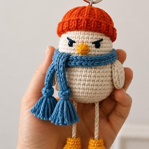 May include: A crocheted bird keychain featuring a cream-colored body, an orange beanie, a blue scarf with tasseled ends, and yellow shoes. The bird has a small orange beak and dark blue embroidered eyes, with a silver metal ring attached at the top.