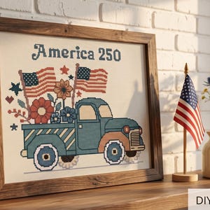 Op de afbeelding: Een ingelijst kruissteek kunstwerk met een blauwe vintage pick-up truck die bloemen en twee Amerikaanse vlaggen vervoert, met de tekst America 250 erboven. Het ontwerp gebruikt rode, witte en blauwe kleuren in een donkerhouten lijst.