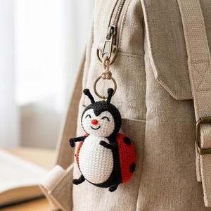 May include: A crocheted ladybug keychain featuring a round white belly, a black head with two antennae, and red wings with black spots. The small stuffed figure has a smiling face with a tiny orange nose and is attached to a metal ring and clasp.