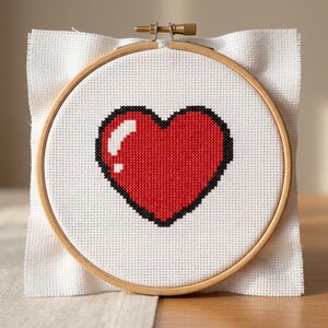 May include: A cross-stitch embroidery of a red heart with a black outline, set in a wooden hoop. The heart has a white highlight, giving it a glossy appearance. The white fabric is visible around the hoop.