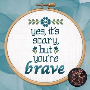 Pu&ograve; includere: Un telaio da ricamo a punto croce con le parole "yes, it's scary, but you're brave" in filo color verde acqua. Il design include accenti floreali e un logo in basso a destra. Il telaio &egrave; su uno sfondo verde acqua e bianco.