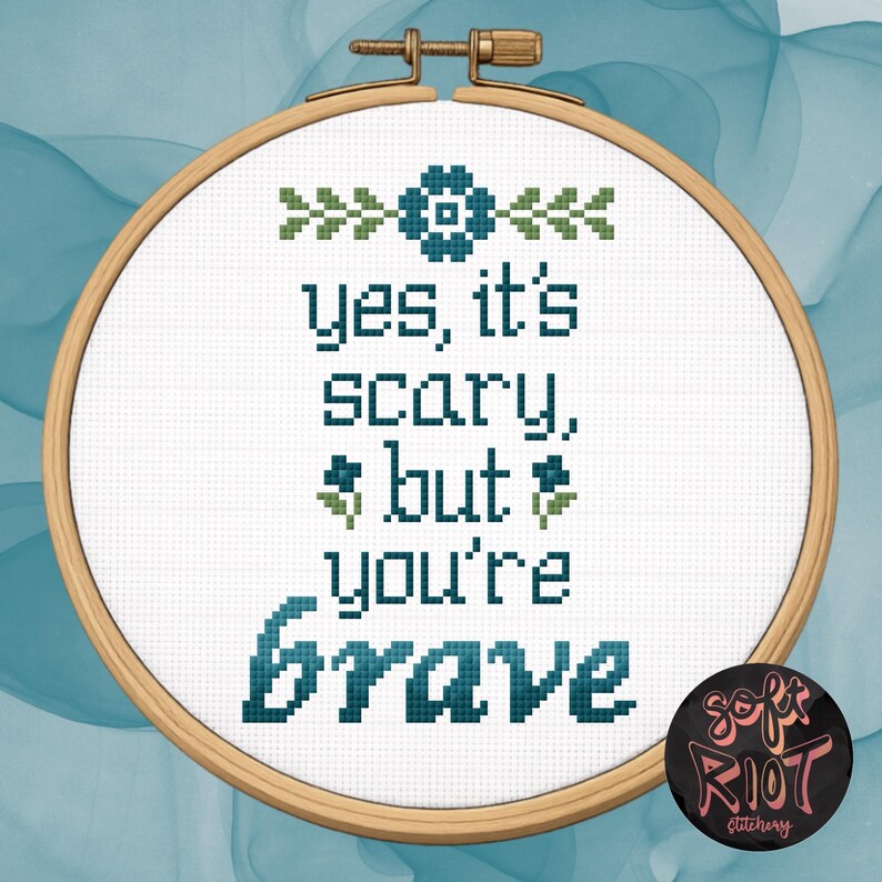 Pu&ograve; includere: Un telaio da ricamo a punto croce con le parole "yes, it's scary, but you're brave" in filo color verde acqua. Il design include accenti floreali e un logo in basso a destra. Il telaio &egrave; su uno sfondo verde acqua e bianco.