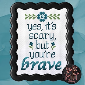 Pu&ograve; includere: Ricamo a punto croce in una cornice nera ondulata. Il testo recita "yes, it's scary, but you're brave" in verde acqua e verde. Una spilla rotonda con la scritta "soft riot stitchery" si trova nell'angolo in basso a destra.
