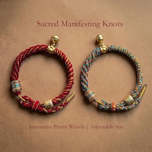 May include: Two interactive prayer wheels with adjustable size. One is red and gold, the other is multicoloured. Each has a gold bell and decorative beads. The text reads "Sacred Manifesting Knots".