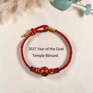 May include: A red braided cord bracelet featuring a central red patterned bead flanked by smaller blue, brown, and gold-accented beads. The bracelet includes a sliding knot closure and text that reads 2027 Year of the Goat Temple Blessed.