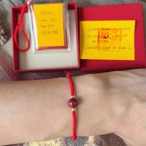 May include: A red string bracelet with a central maroon bead and gold accents. The bracelet is displayed on a wrist, with a red box and yellow cards in the background. The cards have handwritten text and a stamp.