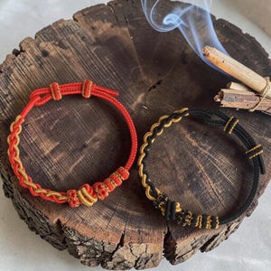 Puede incluir: Dos pulseras hechas a mano, una roja y dorada, la otra negra y dorada. Ambas presentan diseños trenzados y nudos decorativos. Se exhiben sobre una superficie de madera rústica, con humo de incienso en el fondo.