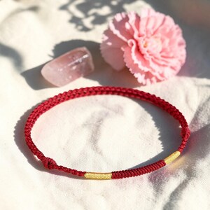 Puede incluir: Una pulsera trenzada roja con un acento dorado, exhibida sobre una superficie blanca. Una flor rosa y un pequeño cristal rosa también están en la imagen. La pulsera es una pieza de joyería simple y elegante.