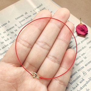 Può includere: Un delicato braccialetto di filo rosso con chiusura dorata è tenuto in una mano. Il braccialetto è circolare e la chiusura è un piccolo design rotondo. Un petalo di rosa rossa secca poggia vicino su una pagina di libro vintage.