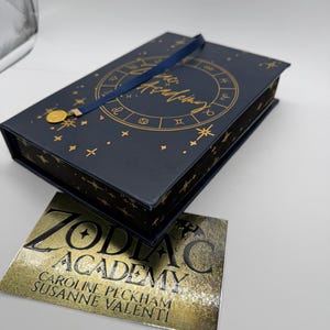 May include: A navy blue book with gold astrological symbols and the words "Zodiac Academy" in gold lettering. A gold medallion hangs from a navy blue ribbon. A gold and silver card with the words "Zodiac Academy" and author names is next to the book.